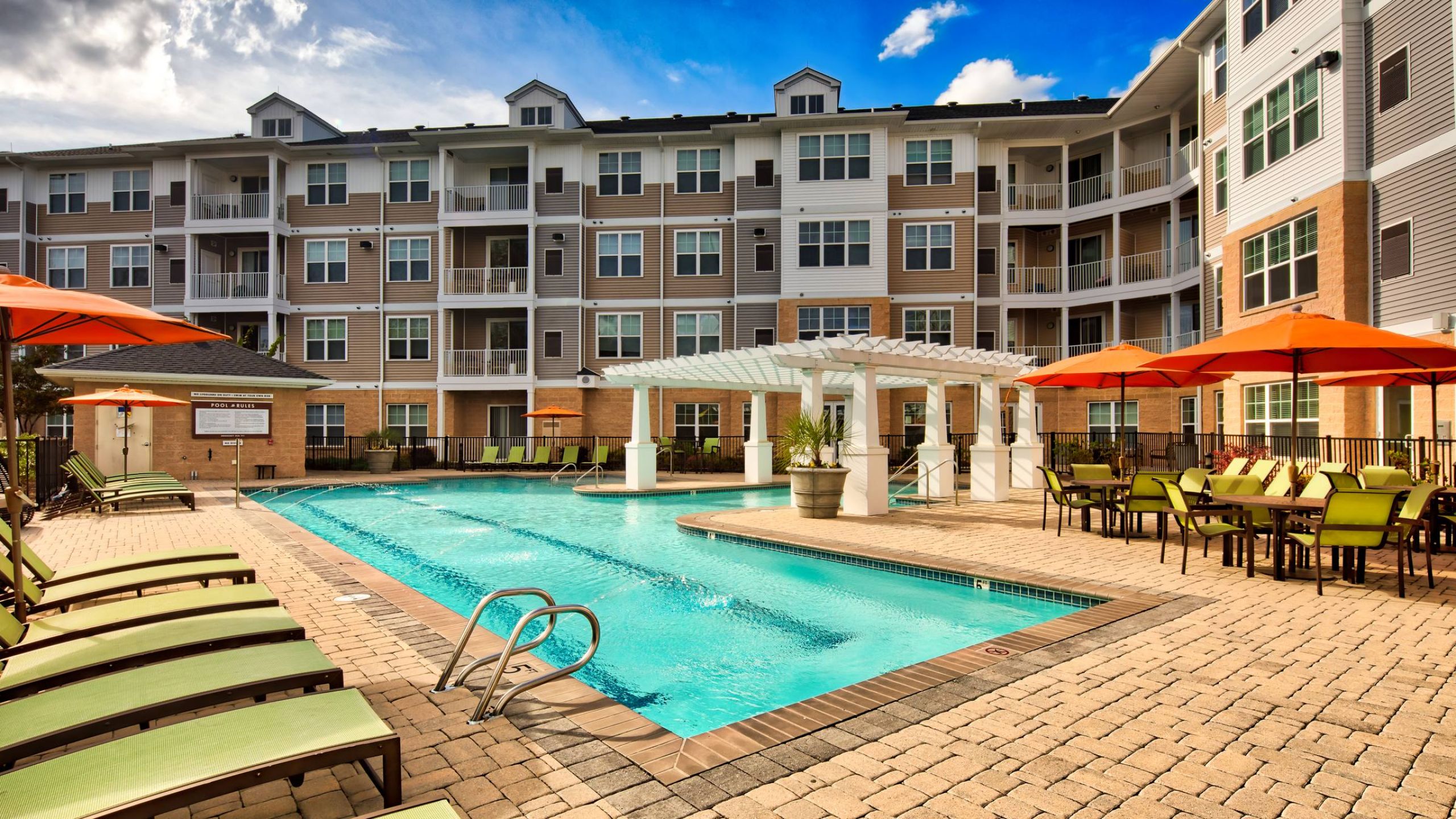 Solace Apartments Outdoor apartment pool area with lounge chairs, tables, and umbrellas in front of a multi-story building.