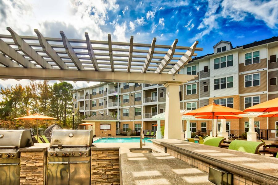 Solace Apartments Outdoor apartment pool area with grills, lounge chairs, orange umbrellas, and a pergola under a blue sky.