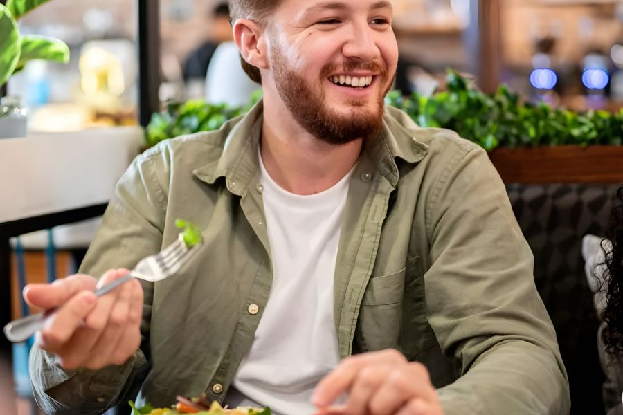 Solace Apartments Smiling man with a beard eating a salad at a restaurant table with plants in the background.