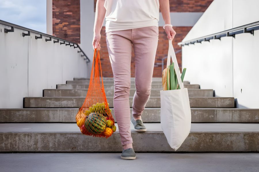 Person walking down stairs carrying reusable bags with groceries and vegetables.