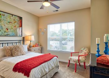 Solace Apartments Cozy bedroom with a bed, red blanket, floral chair, large window, and colorful wall art.