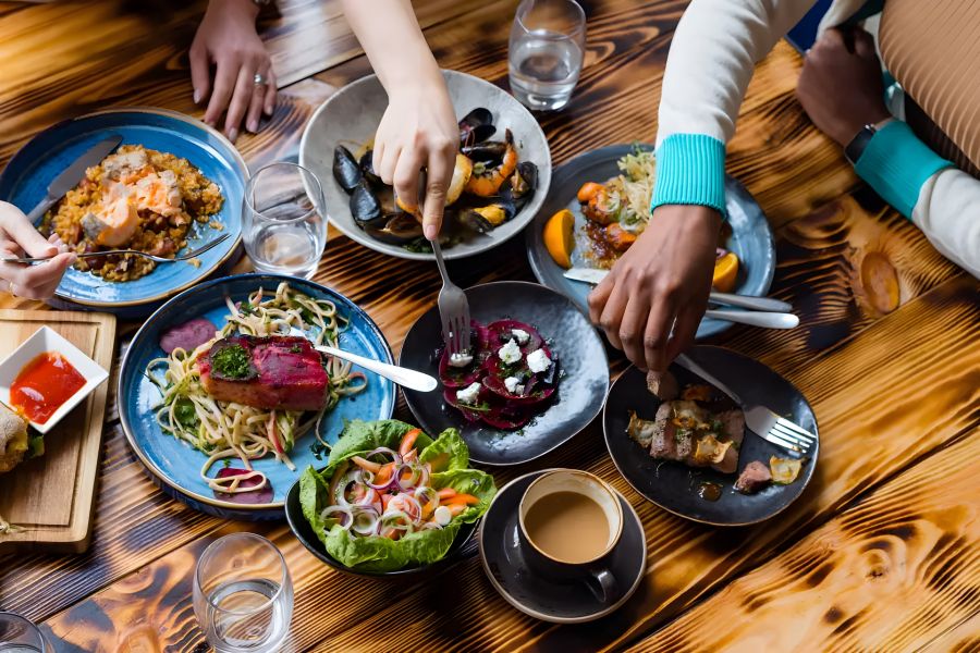 Solace Apartments A group of people share assorted dishes at a wooden table, reaching for food and enjoying a meal together.