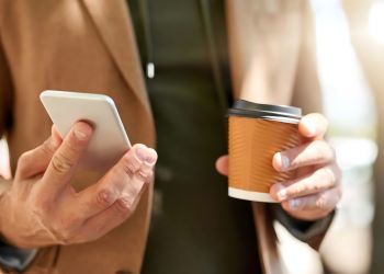 Solace Apartments A person holds a smartphone in one hand and a takeaway coffee cup in the other, outdoors.