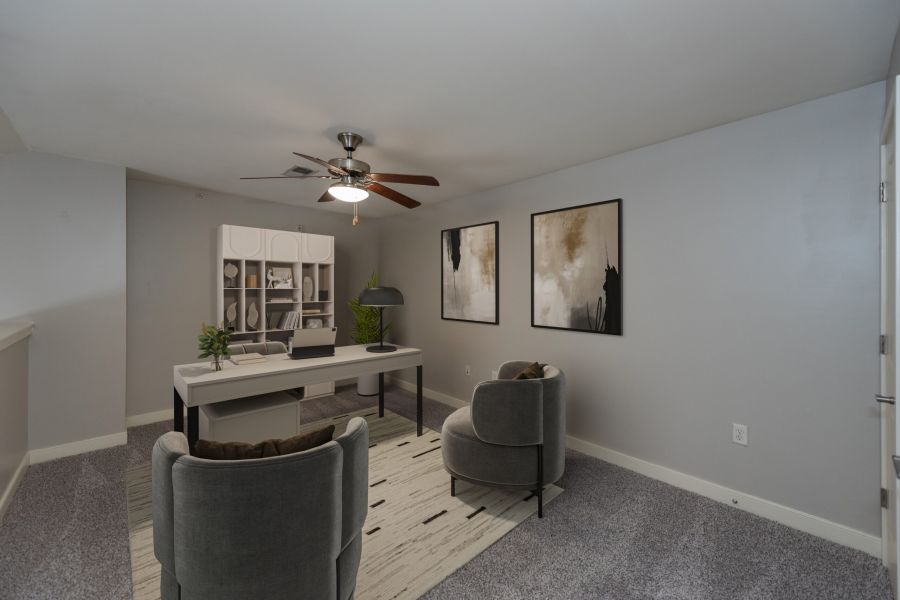Modern home office with a desk, two chairs, wall art, carpet, and a ceiling fan in a gray-toned room.